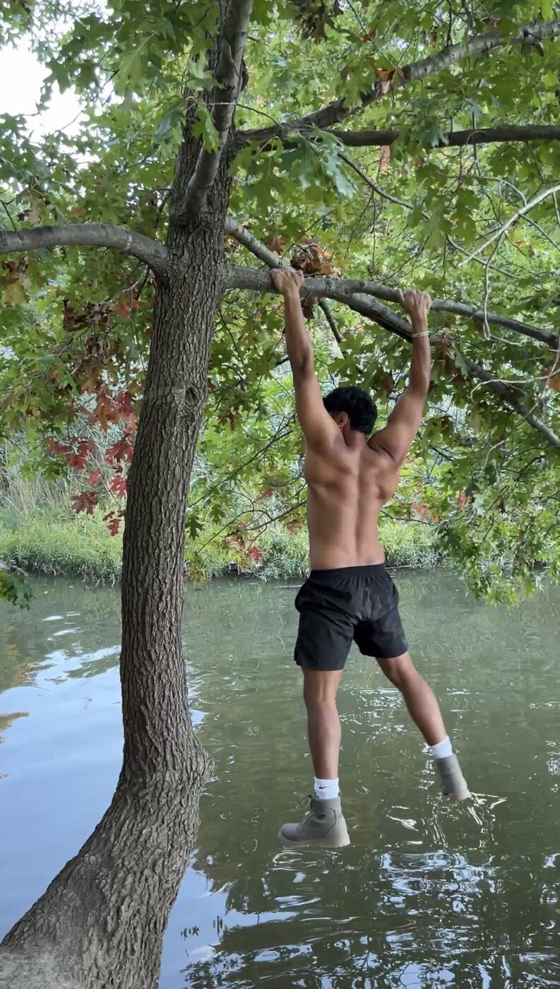 Akshat doing pull-ups on a tree branch over a river.